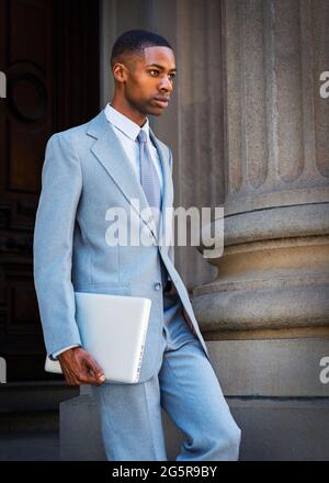 Ein junger afroamerikanischer Geschäftsmann, der einen hellgrauen Anzug, eine weiße Unterhemd, eine Krawatte und einen Laptop trägt, geht vom Wintag herunter Stockfoto