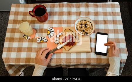Zenitale Aufnahme der Hände einer Latina-Frau, Scrollen auf ihrem Handy beim Frühstück. Platz kopieren Stockfoto