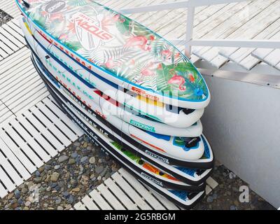 Russland, Sotschi 06.06.2021. Helle mehrfarbige Surfbretter, die in der Nähe von Treppen am Kiesstrand mit hölzernen Gehwegen gestapelt sind Stockfoto