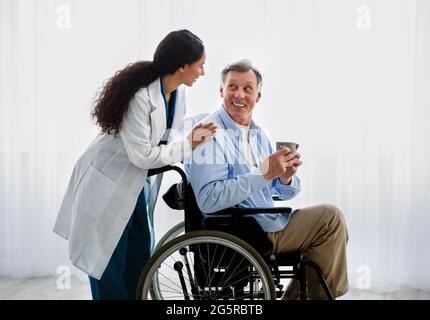 Junge Ärztin, die älteren behinderten Menschen im Rollstuhl hilft und sich im Altersheim um ältere Patienten kümmert Stockfoto