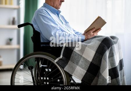 Beschnittene Ansicht eines behinderten älteren Mannes im Rollstuhl, der ein Buch im Gebäude liest Stockfoto