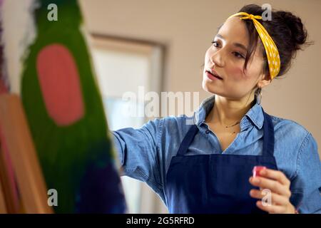 Portrait einer jungen Frau in Schürze, die inspiriert aussieht, während sie in einem gemütlichen Workshop an abstrakter Malerei arbeitet Stockfoto