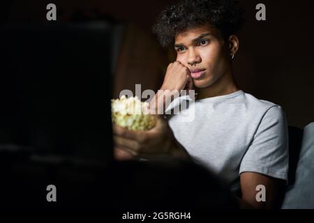 Langweiliger Film. Junger Mann, der sich beim Ansehen eines Films gelangweilt und Popcornschüssel in der Hand hält und zu Hause auf einem Sofa im dunklen Zimmer sitzt Stockfoto