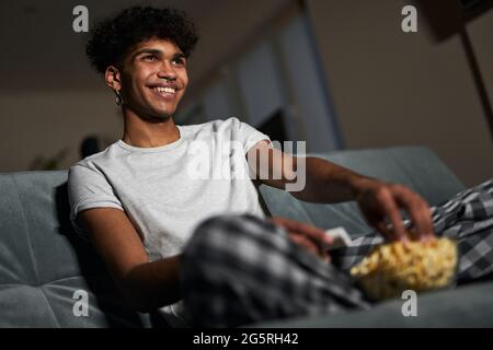 Fröhlicher junger Mann im Schlafanzug, der Popcorn aus einer Schüssel nimmt, während er sich eine Komödie anschaut und zu Hause auf einer Couch sitzt Stockfoto