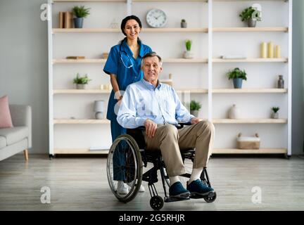 In voller Länge Porträt eines älteren behinderten Mannes im Rollstuhl und seines jungen Betreuers, der im Altersheim vor der Kamera lächelt Stockfoto