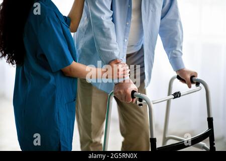 Beschnittene Ansicht eines älteren Mannes, der Gehhilfe, eine junge Krankenschwester, die ihn im Altersheim unterstützt und unterstützt, in Nahaufnahme verwendet Stockfoto