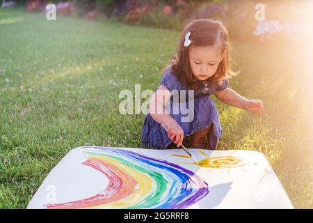 Kleines Mädchen 2-4 Jahre alt malt Regenbogen und Sonne auf ein großes Blatt Papier sitzen auf grünem Rasen in Sonnenlicht Stockfoto