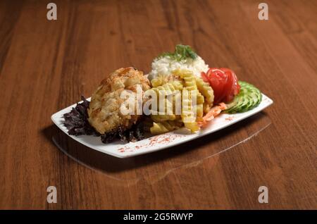 Hühnerschnitzel in Kiew panierte in Semmelbröseln, mit Reis, Pommes frites, Tomaten und Gurken auf einem weißen Teller auf dem Tisch Stockfoto