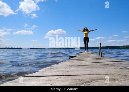 Mädchen, das am Pier in der Nähe des Flusses steht, ihre Hände nach oben hebt, genießt das sonnige Wetter. Stockfoto