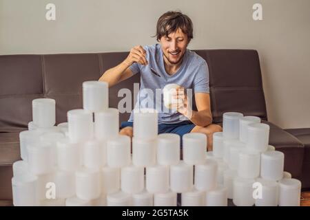 Der Mann isst Joghurt und ist von vielen Joghurtgläsern umgeben, die er in einem Jahr verwendet. Eine riesige Menge von Plastikbehältern für Lebensmittel. Übermäßiger Verbrauch von Stockfoto