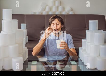 Der Mann isst Joghurt und ist von vielen Joghurtgläsern umgeben, die er in einem Jahr verwendet. Eine riesige Menge von Plastikbehältern für Lebensmittel. Übermäßiger Verbrauch von Stockfoto