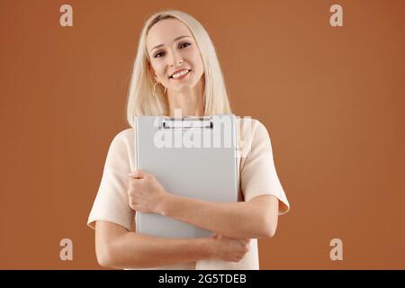 Porträt einer positiven erfolgreichen jungen kaukasischen Frau umarmt Zwischenablage vor braunem Hintergrund Stockfoto