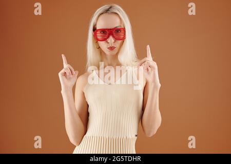 Coole junge kaukasische Frau in rot gestreifter Brille, die Lippen puckert und die Finger vor braunem Hintergrund zeigt Stockfoto