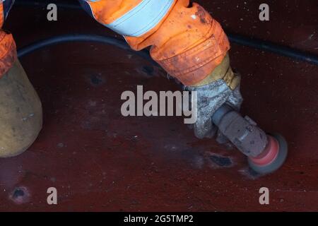 Arbeiter Hand gekleidet in orange Gesamt- und Schutzhandschuh Betrieb Luft pneumatische Winkelschleifer, um den Rost von der Metalloberfläche zu entfernen. Stockfoto