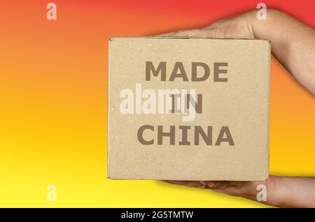 Hände, die einen Karton auf einem farbigen Hintergrund halten. Ein Mann mit einem quadratischen Paket aus recyceltem Material. Inschrift hergestellt in China. Globale Handelskonce Stockfoto