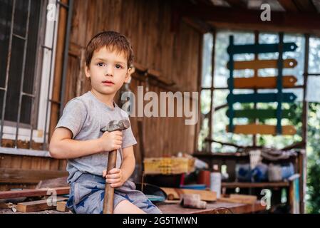 Der kleine Junge hämmert in der Tischlerwerkstatt mit einem Hammer Nägel in ein Holzbrett Stockfoto