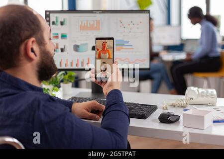 Behinderter, ungültiger Manager, der während der Arbeitszeit mit Freunden, die das Smartphone halten, auf Videocall spricht. Im Rollstuhl sitzender Mitarbeiter, der im Finanzbüro arbeitet. Stockfoto