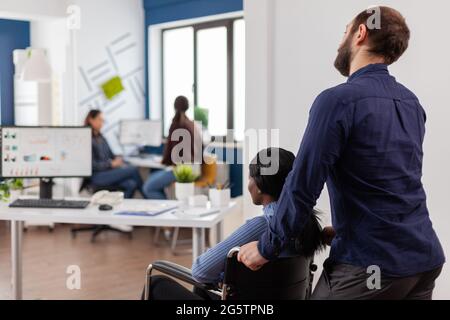 Ein Angestellter hilft seinem ungültigen afrikanischen Kollegen, der am Arbeitsplatz mit dem Rollstuhl in die Nähe des Schreibtisches kommt. Schwarze gelähmte Frau begrüßt Kollegen, die bei einem Finanzprojekt arbeiten. Stockfoto