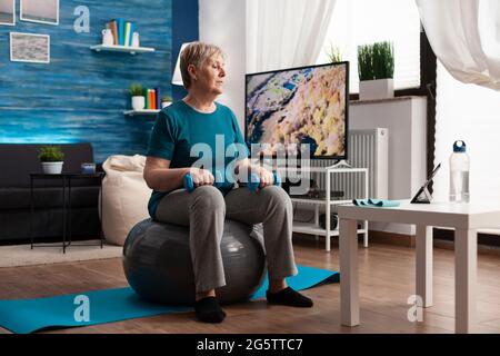 Fokussierte ältere Frau beim Armtraining mit Workout-Hanteln, die auf schweizer Ball im Wohnzimmer sitzen. Rentner in Sportbekleidung, die Online-Aerobic-Training auf Tablet Ausübung Körperwiderstand Stockfoto