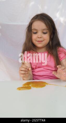 Kind spielt handgemachtes Spielzeug namens Slime. Nettes Mädchen in einem rosa T-Shirt spielt mit einem gelben Schleim. Zeitgenössische Kreativität. Stretching, scaming, Modellierung Stockfoto