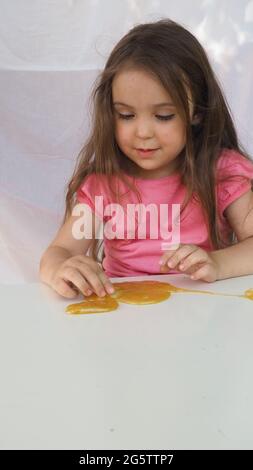 Kind spielt handgemachtes Spielzeug namens Slime. Nettes Mädchen in einem rosa T-Shirt spielt mit einem gelben Schleim. Zeitgenössische Kreativität. Stretching, scaming, Modellierung Stockfoto