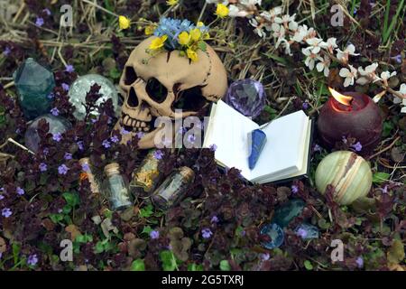 Stillleben mit Totenkopf, Blumen, Kristallen und Hexenflaschen mit Tränke und Kräutern draußen. Esoterischer, gotischer und okkulter Hintergrund, Halloween Mystiker und Stockfoto