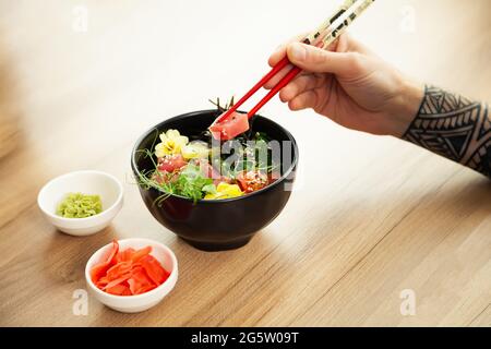Ein Mann isst einen Poke-Salat mit Thunfisch in einer Schüssel mit Essstäbchen. Den Thunfischsalat in eine Schüssel geben. Junger Mann in einem Restaurant, der einen Sack-Salat isst. Asiatisch Stockfoto