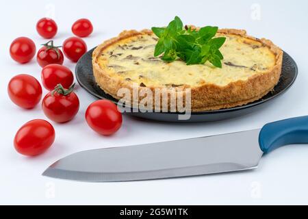 Nahaufnahme großer Studioaufnahme von frisch gebackenem gelbem französischem Salzkuchen oder Quiche, mit Pilzen, roten Kirschtomaten und grüner Minze, auf schwarzem Design pla Stockfoto