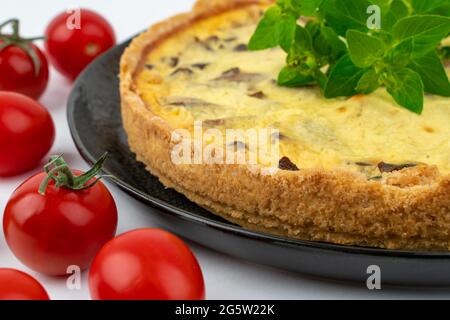 Nahaufnahme große Studioaufnahme von frisch gebackenen gelben französischen salzigen Kuchen oder Quiche, mit Pilzen, roten Kirschtomaten, auf einem schwarzen Design-Teller, isoliert Stockfoto