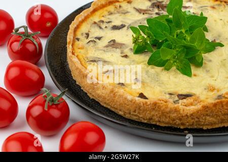 Nahaufnahme große Studioaufnahme von frisch gebackenen gelben französischen salzigen Kuchen oder Quiche, mit Pilzen, roten Kirschtomaten, auf einem schwarzen Design-Teller, isoliert Stockfoto