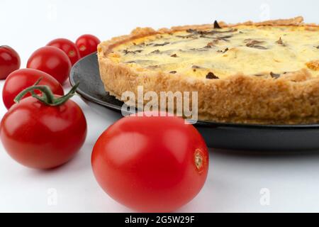 Nahaufnahme große Studioaufnahme von frisch gebackenen gelben französischen salzigen Kuchen oder Quiche, mit Pilzen, roten Kirschtomaten, auf einem schwarzen Design-Teller, isoliert Stockfoto