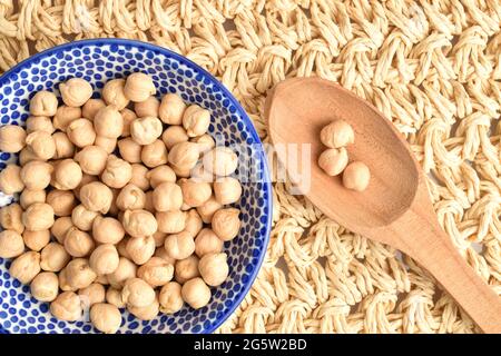 Bio-natürliche, ungekochte Kichererbsen in einer keramischen Untertasse mit Holzlöffel, Nahaufnahme, auf einer Strohmatte, Draufsicht. Stockfoto
