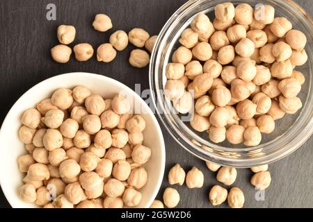 Natürliche, ungekochte Kichererbsen aus biologischem Anbau in Glas- und Keramikgeschirr, Nahaufnahme, auf Schiefertafel, Draufsicht. Stockfoto