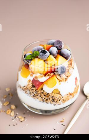 Griechischer Joghurt, Müsli und Früchte im Glas. Gesundes vegetarisches Frühstücksparfait mit Pfirsich, Heidelbeeren, Haferhoniggranola und Joghurt, beigefarbener Hintergrund Stockfoto