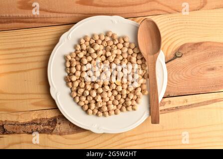 Natürliche, ungekochte Kichererbsen aus biologischem Anbau in einer weißen Keramikplatte mit Holzlöffel, Nahaufnahme, auf einem Holztisch, Draufsicht. Stockfoto