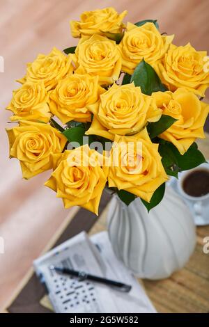 Gelbe Rosen mit Kuskuhrblatt in einer weißen Vase auf einem kleinen Couchtisch mit einem Kreuzworträtsel einer Zeitung, einem Stift und einem Kaffee in einem Zimmer Stockfoto