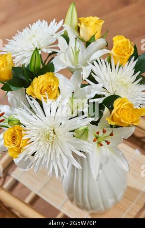 Eine Blumenvase mit gelben Rosen, weißem Spray Anastasia und weißen asiatischen Lilien und Ruscus Leaf auf einem Bambustisch drinnen Stockfoto