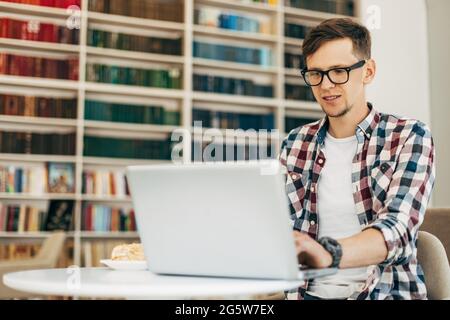 Junger lächelnder Geschäftsmann, benutzt Laptop, Mann arbeitet im Internet sitzt an einem Tisch mit einem modernen Laptop-Computer in einem Café vor dem Hintergrund o Stockfoto