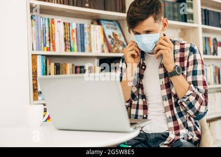 Der junge Student trägt in einem karierten Casual-Hemd eine medizinische Schutzmaske zum Schutz vor Coronaviren, der Student arbeitet an einem Laptop im unive Stockfoto