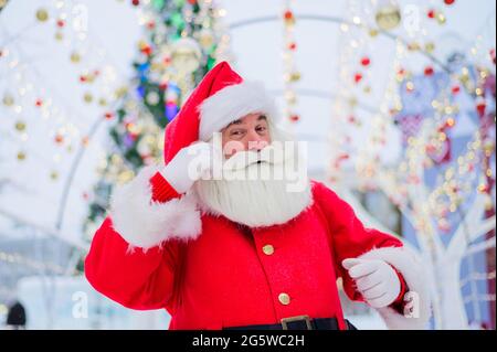 Porträt eines älteren Mannes, der als weihnachtsmann gekleidet ist, auf dem Hintergrund eines weihnachtsbaums im Freien. Stockfoto