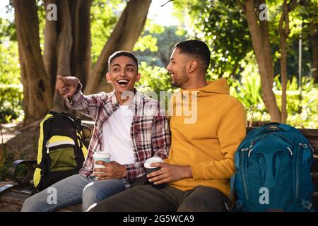 Zwei glückliche gemischte Rennfreunde, die mit Rucksäcken auf der Parkbank sitzen und sich unterhalten Stockfoto