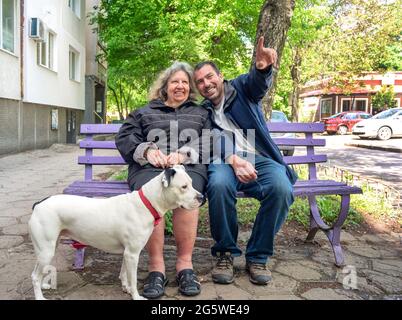 Generationen Ältere Frau und Sohn mit Hund sitzen auf einer Bank im Freien lächelnd Stockfoto