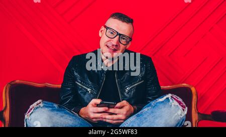 Hübscher junger Mann mit Brille, die mit Smartphone auf einem kleinen Sofa an einer roten Wand ruht. Stockfoto