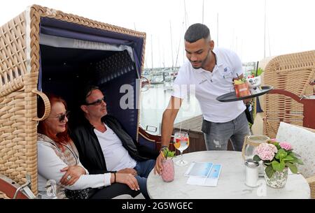 30. Juni 2021, Mecklenburg-Vorpommern, Kühlungsborn: Auf der Terrasse des Restaurants Vielmeer am Bootshafen serviert Ibrahim Hussein Gäste, die in einem Strandstuhl sitzen. Der Neubeginn von Gastronomie und Tourismus nach monatelanger Corona-Zwangspause hat dem Arbeitsmarkt in Mecklenburg-Vorpommern einen starken Schub gegeben. Für den Monat Juni meldete die Nord-Regionaldirektion der Bundesagentur für Arbeit in Kiel am Mittwoch bundesweit 61,600 Arbeitslose. Das waren 4800 weniger als im Vormonat Mai und 4300 weniger als im Juni 2020. Foto: Bernd Wüstneck/dpa-Zentralbild/dpa Stockfoto