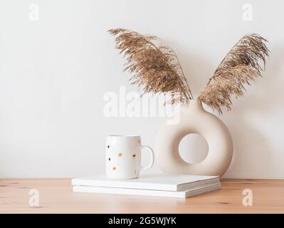 Gemütliches Herbsthaus Stillleben von trockenem Schilf Zweige in Vase, weißer Keramik-Becher mit goldenen Sternen und Zeitschriften auf Holztisch. Vorderansicht. Stockfoto