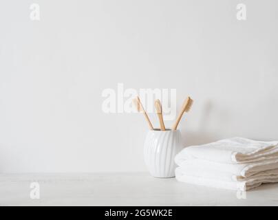 Minimalistischer Badehintergrund mit Bambuszahnbürsten aus Keramikglas und weißen Handtüchern auf weißem Hintergrund. Vorderansicht. Speicherplatz kopieren Stockfoto