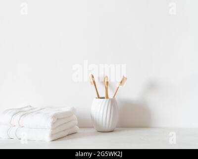 Minimalistischer Badehintergrund mit Bambuszahnbürsten aus Keramikglas und weißen Handtüchern auf weißem Hintergrund. Vorderansicht. Speicherplatz kopieren Stockfoto