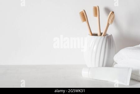 Minimalistischer Badehintergrund mit Bambuszahnbürsten aus Keramikglas, weißer Zahnpasta-Tube und weißen Handtüchern. Vorderansicht. Speicherplatz kopieren Stockfoto