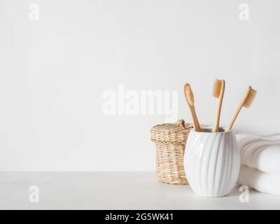 Minimalistischer Badehintergrund mit Bambuszahnbürsten aus Keramikglas, Korbbox und weißen Handtüchern auf weißem Hintergrund. Vorderansicht. Speicherplatz kopieren Stockfoto