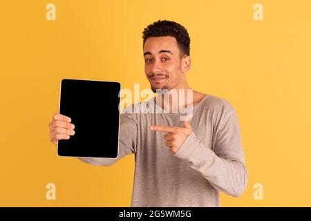 Zeigt auf das Tablett. Kaukasischer Mann Porträt isoliert auf gelbem Hintergrund mit Copyspace. Gutaussehende männliche Modell im Street-Stil. Stockfoto